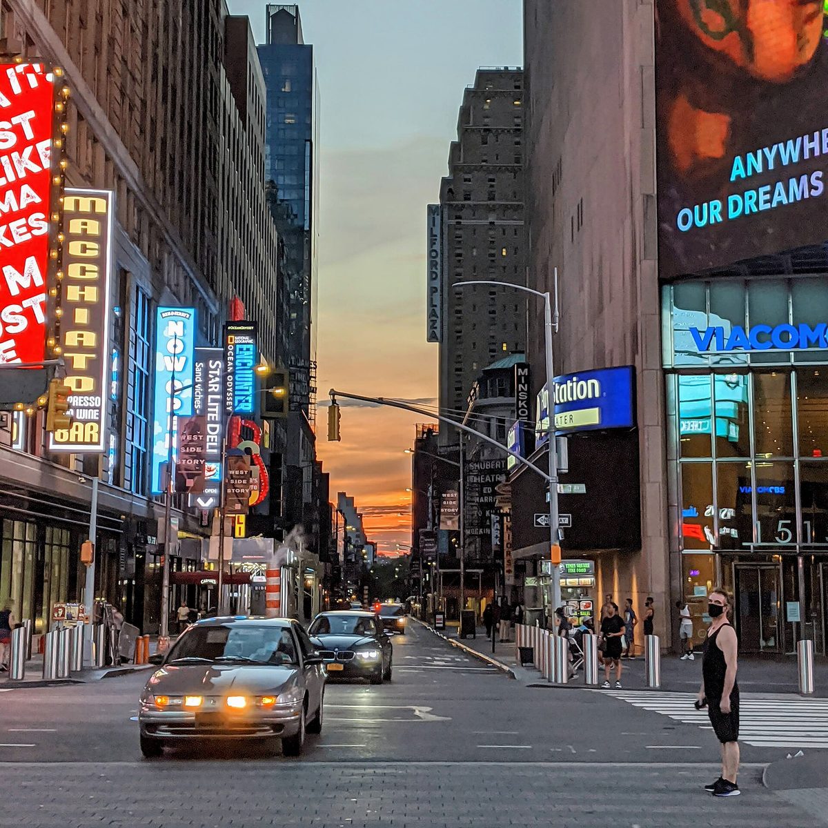 A New York street corner at sunset near Times Square, with lit delicatessen and theatre signs, cars waiting at a crossing and orange sky between the buildings.