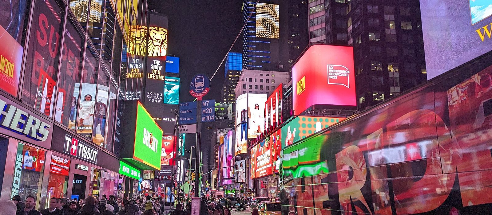 Illuminated advertising screens and signs crowding the buildings of Times Square in New York at dusk, with pedestrians filling the plaza below.