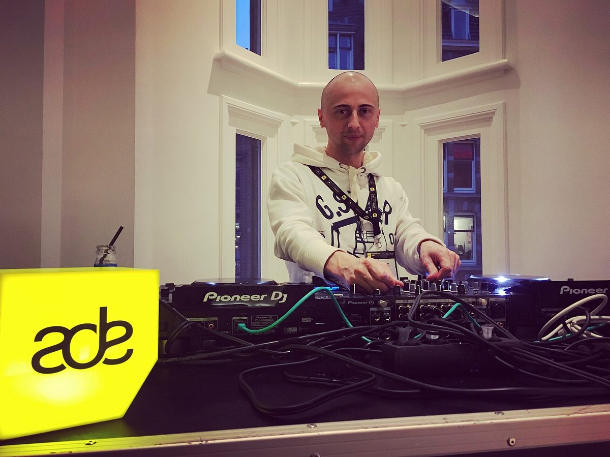 A DJ playing on Pioneer DJ equipment at an Amsterdam Dance Event booth, with a bright yellow ADE logo panel mounted on the front of the table.