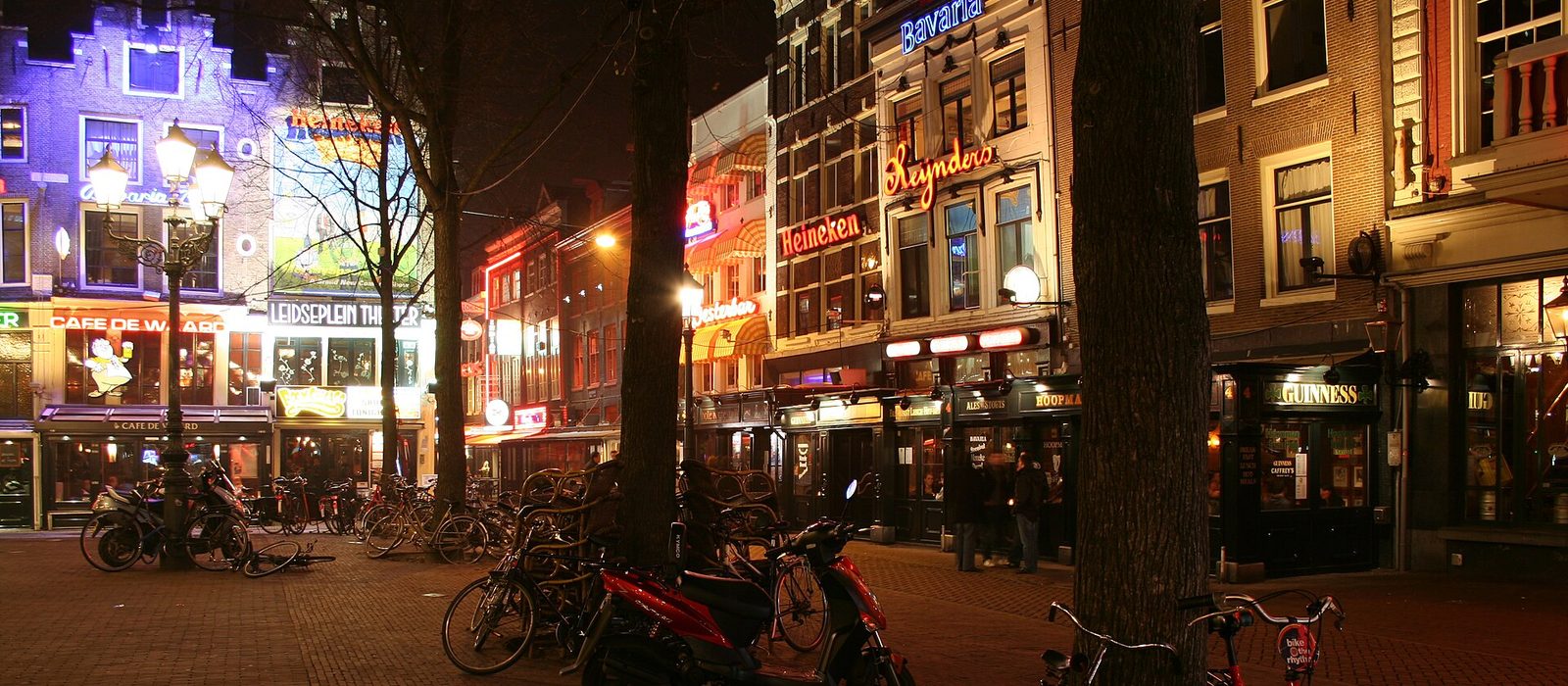 Leidseplein in Amsterdam at night, lined with lit bar and cafe signs including Heineken and Bavaria, with bicycles and scooters parked along the brick square.