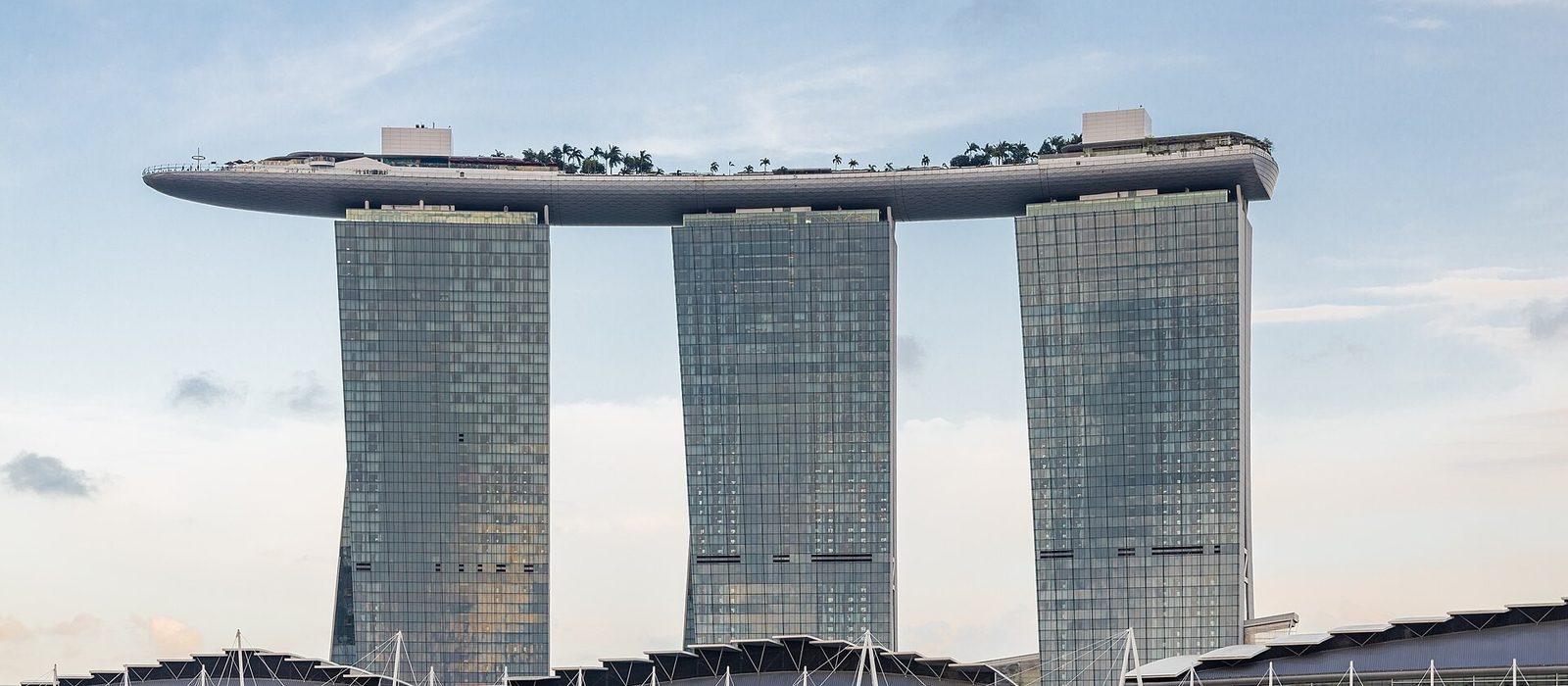 The three towers of Marina Bay Sands in Singapore rising above the waterfront, joined at the top by the boat-shaped SkyPark, under a pale blue sky.