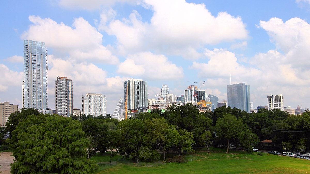 The downtown Austin skyline of glass towers and cranes seen across a green park under a bright cloudy sky.