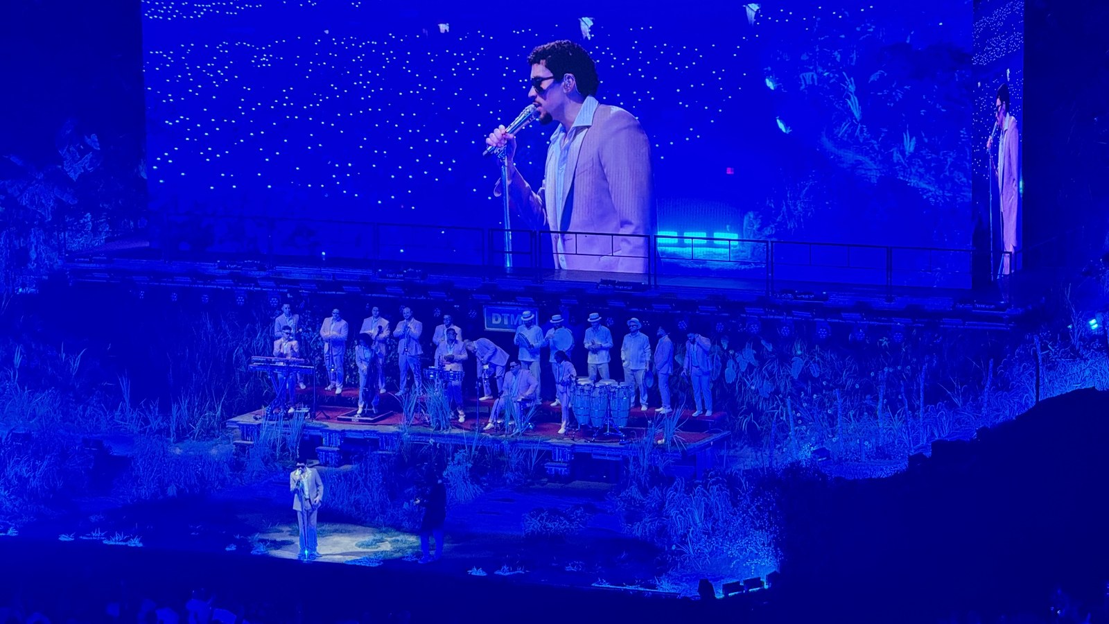 Bad Bunny performing on a stadium stage with a live band, shown on the venue's large video screen