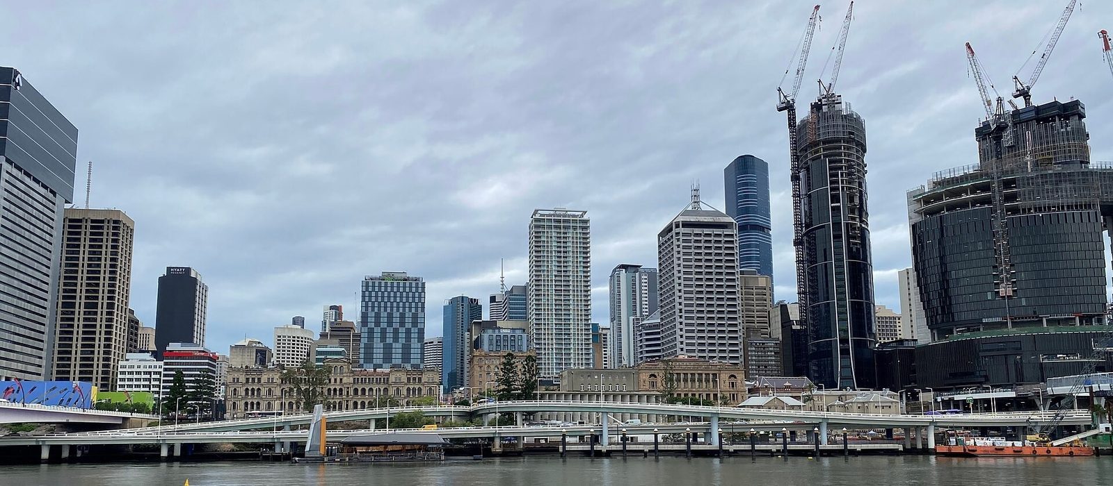 The Brisbane central business district seen across the Brisbane River, a row of glass towers and construction cranes above the riverside walkway.