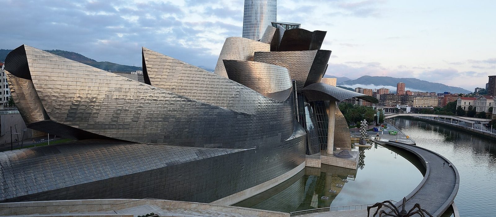 The Guggenheim Museum Bilbao seen from across the Nervion river, its curved titanium panels catching the light, with Louise Bourgeois's steel spider sculpture standing on the terrace beside it.