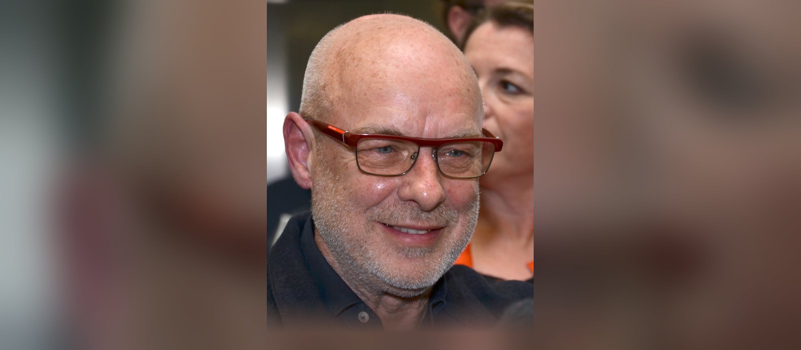 Brian Eno smiling in red-framed glasses at an event in Prague in 2017