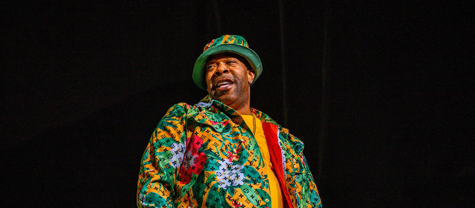 Busta Rhymes performing on stage in a patterned jacket and bucket hat against a dark backdrop