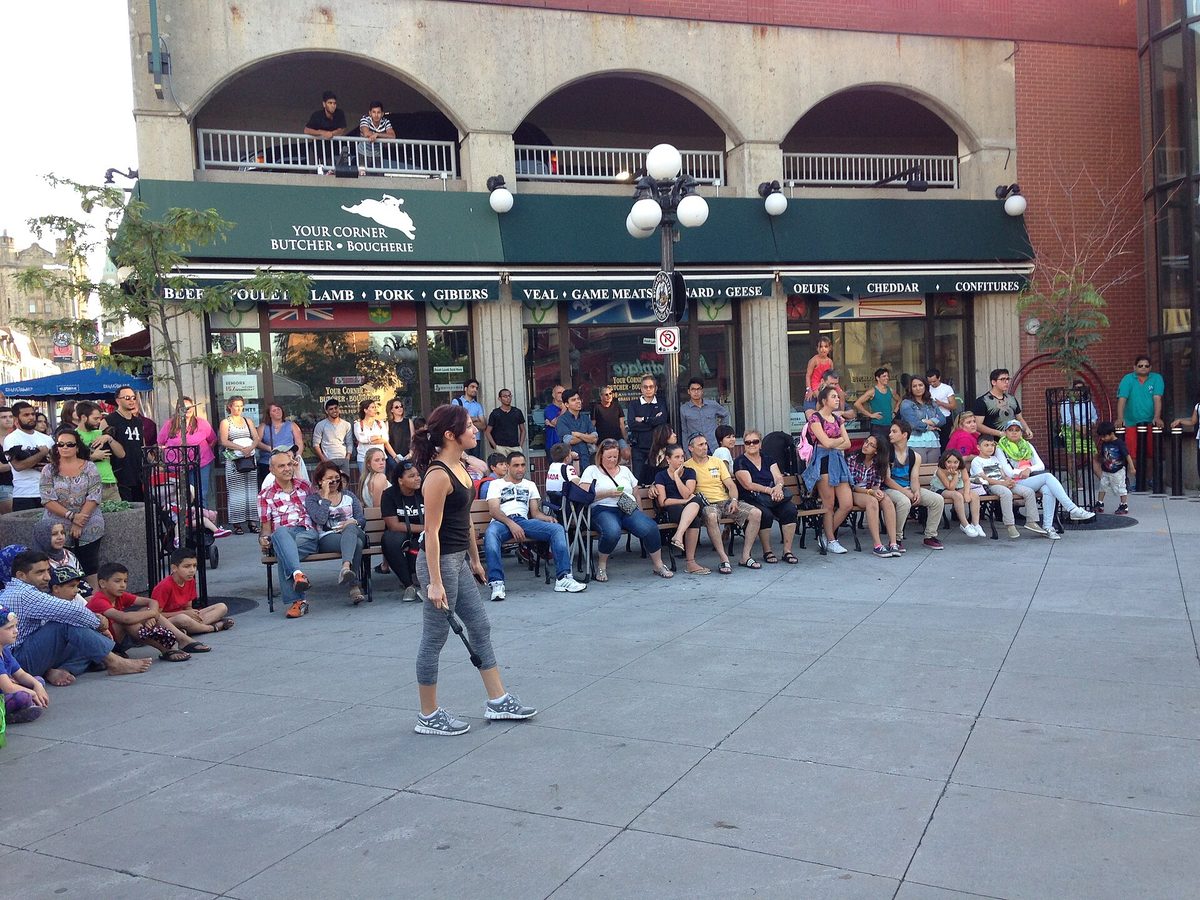 A crowd seated and standing along a paved street in Ottawa's ByWard Market watching a street performance, with market shopfronts and awnings behind them.
