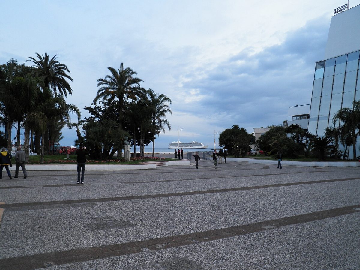 The open plaza beside the Palais des Festivals in Cannes, lined with tall palm trees, with people crossing towards the seafront.