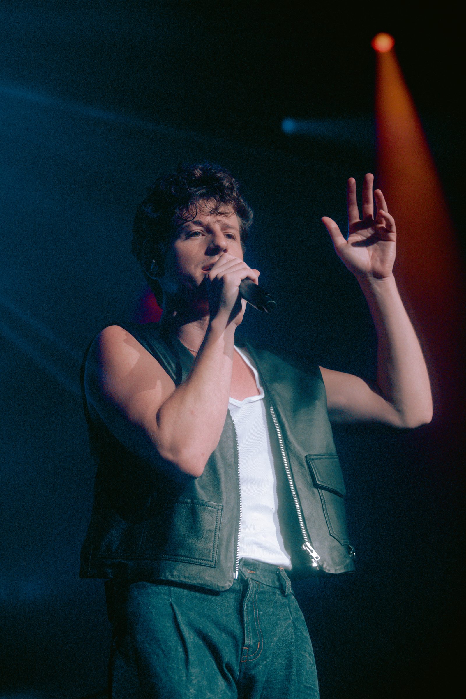 Charlie Puth singing into a microphone on stage in a leather vest