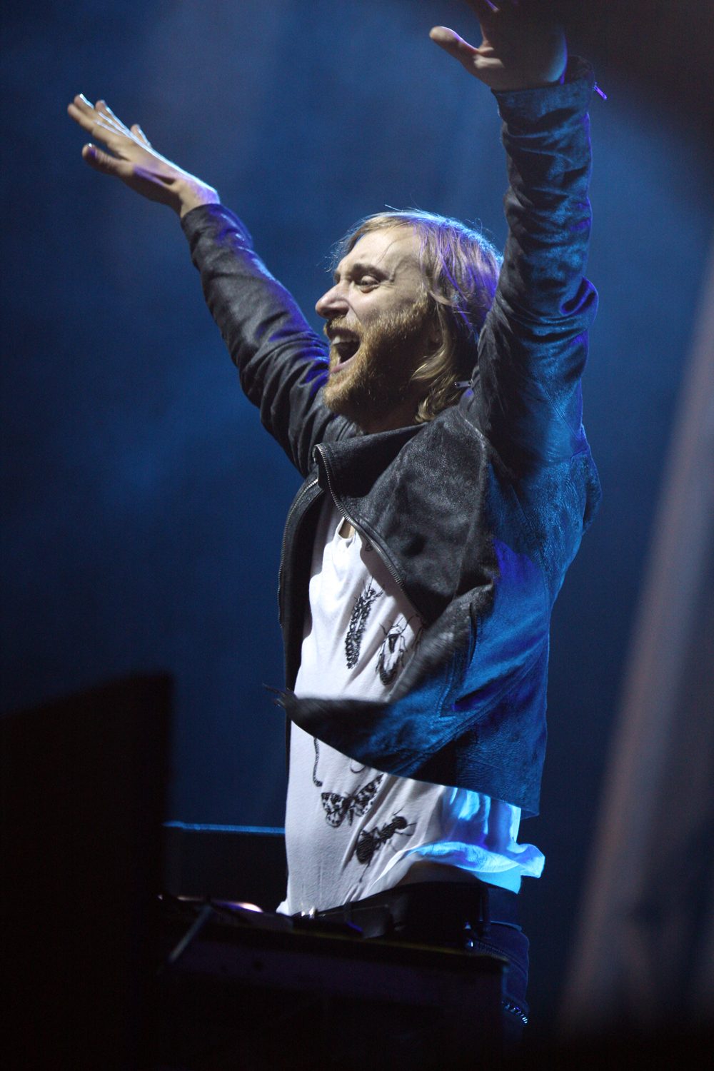 David Guetta with both arms raised on stage under blue lights