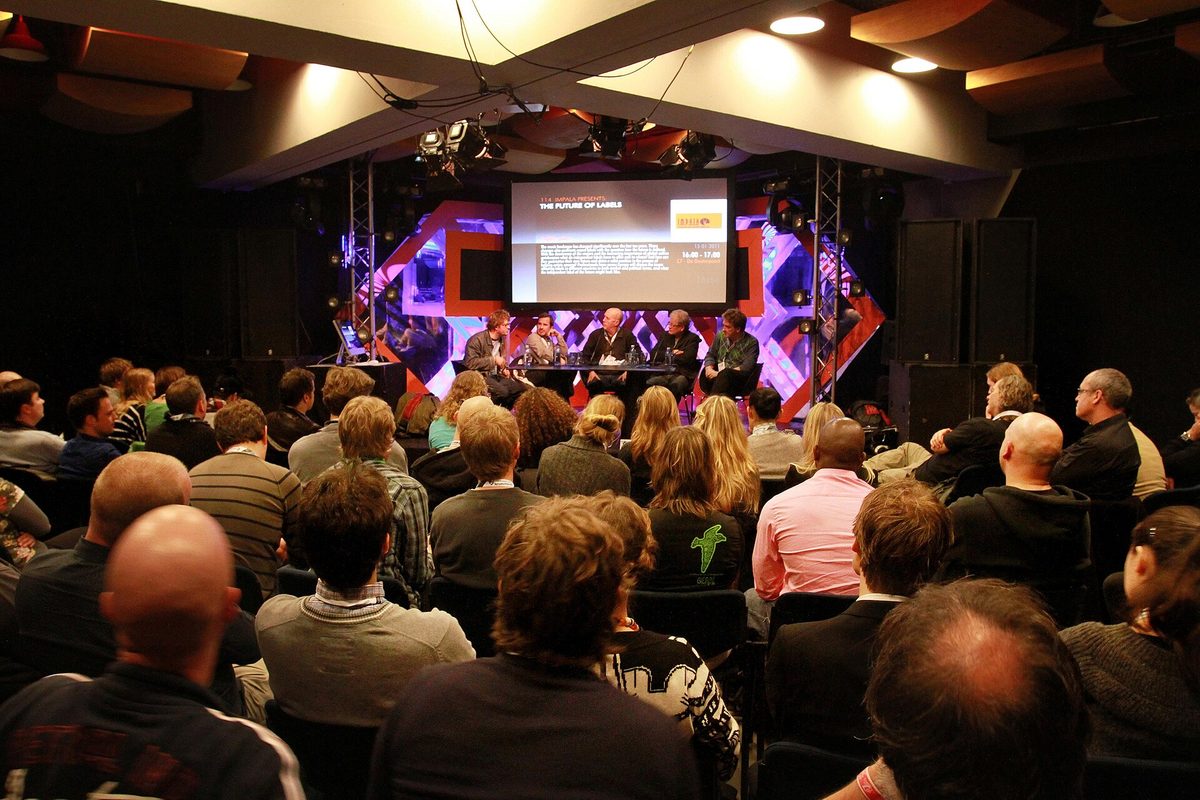 A packed conference audience seen from behind, facing a lit panel stage with a presentation screen at the ESNS conference.