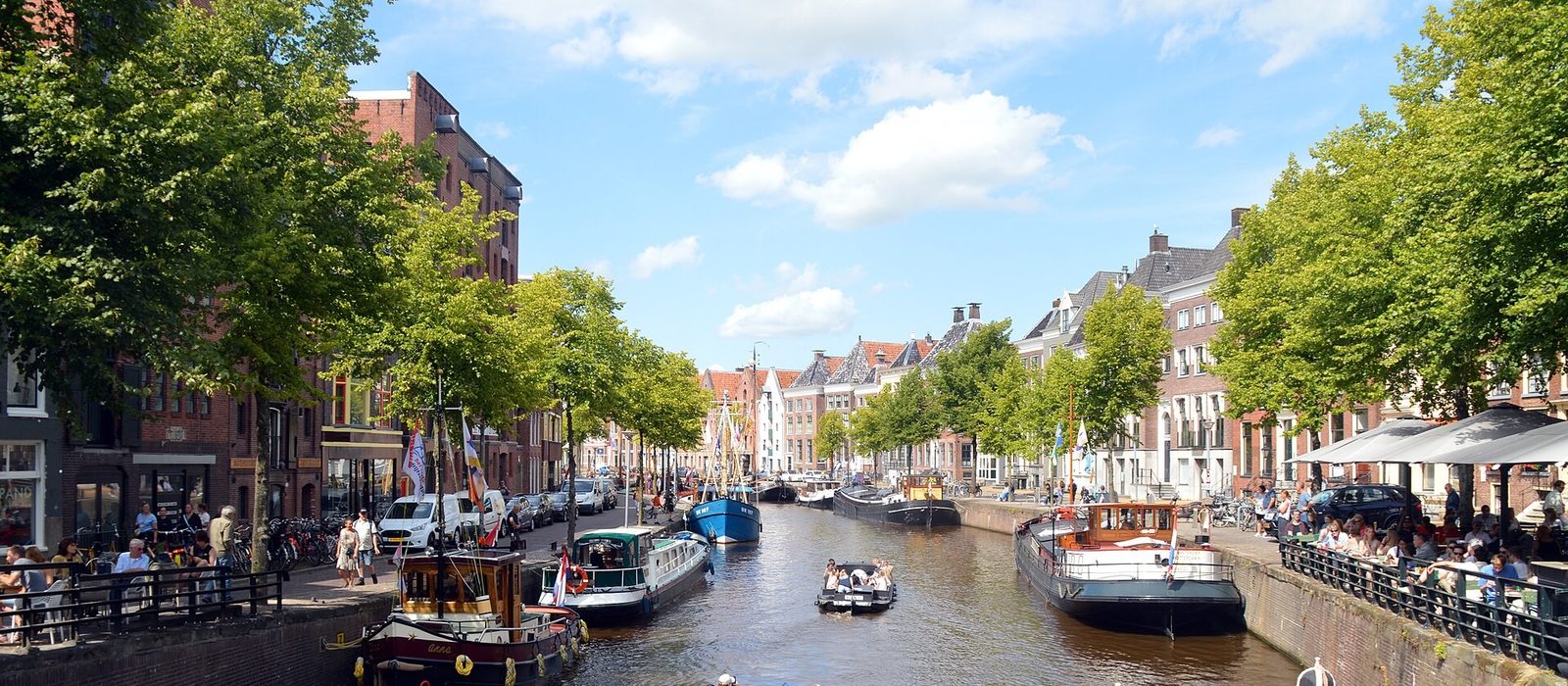 A canal in central Groningen lined with gabled houses and plane trees, with moored barges, a small tour boat on the water and cafe terraces along the quay.