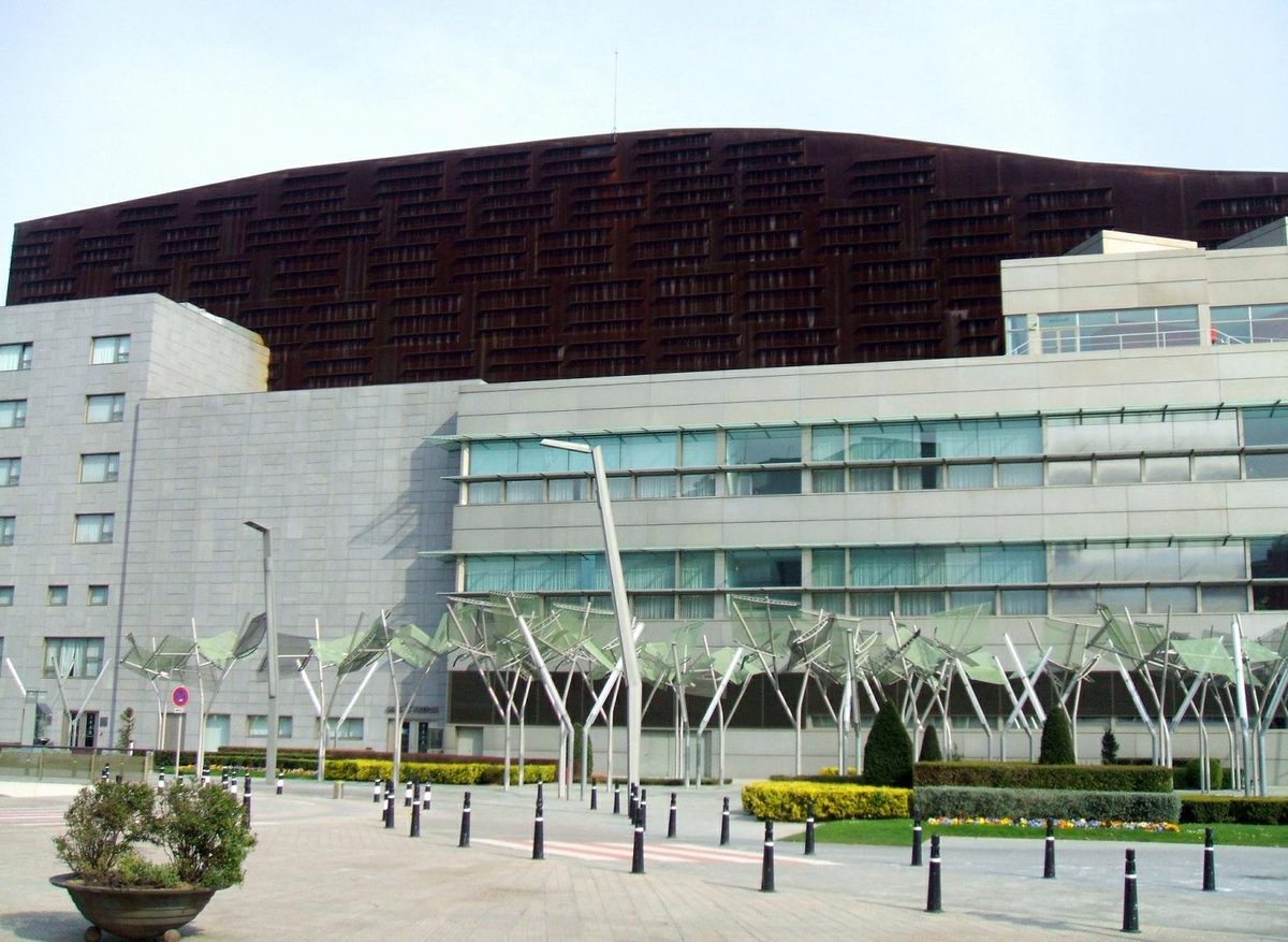 The full facade of the Euskalduna Palace in Bilbao seen from the street, showing its curved rust-coloured upper storeys above glass and stone lower floors.