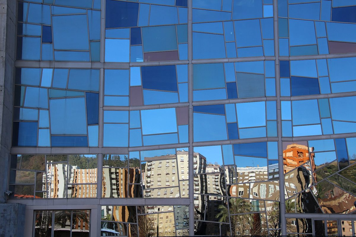 A grid of blue and clear glass panels on the Euskalduna Palace reflecting the surrounding Bilbao buildings and hillside.