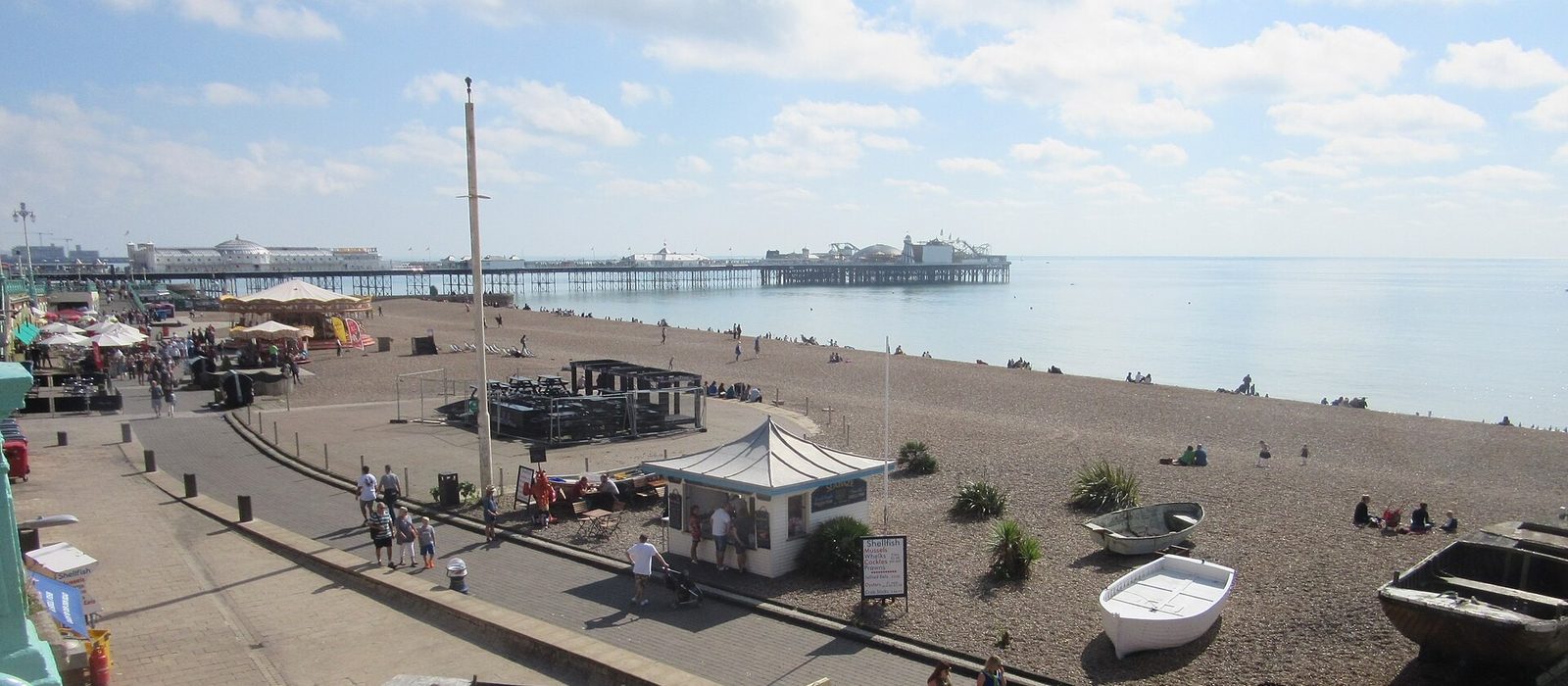 Brighton beach and promenade on a bright day, with the Palace Pier stretching out over the sea in the distance and wooden fishing boats drawn up on the shingle.