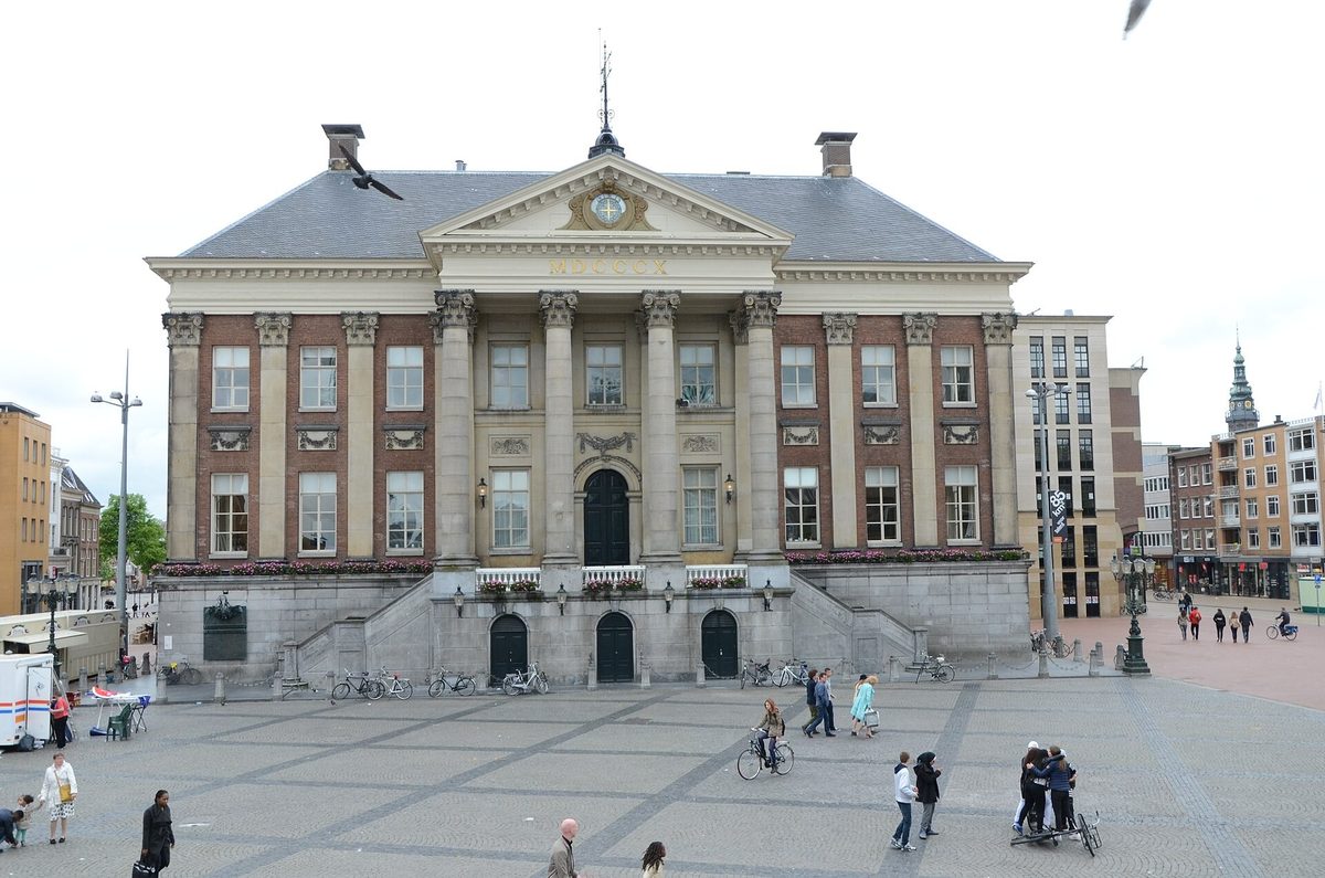 The neoclassical old town hall on the Grote Markt in Groningen, with people and cyclists crossing the open brick-paved square in front of it.