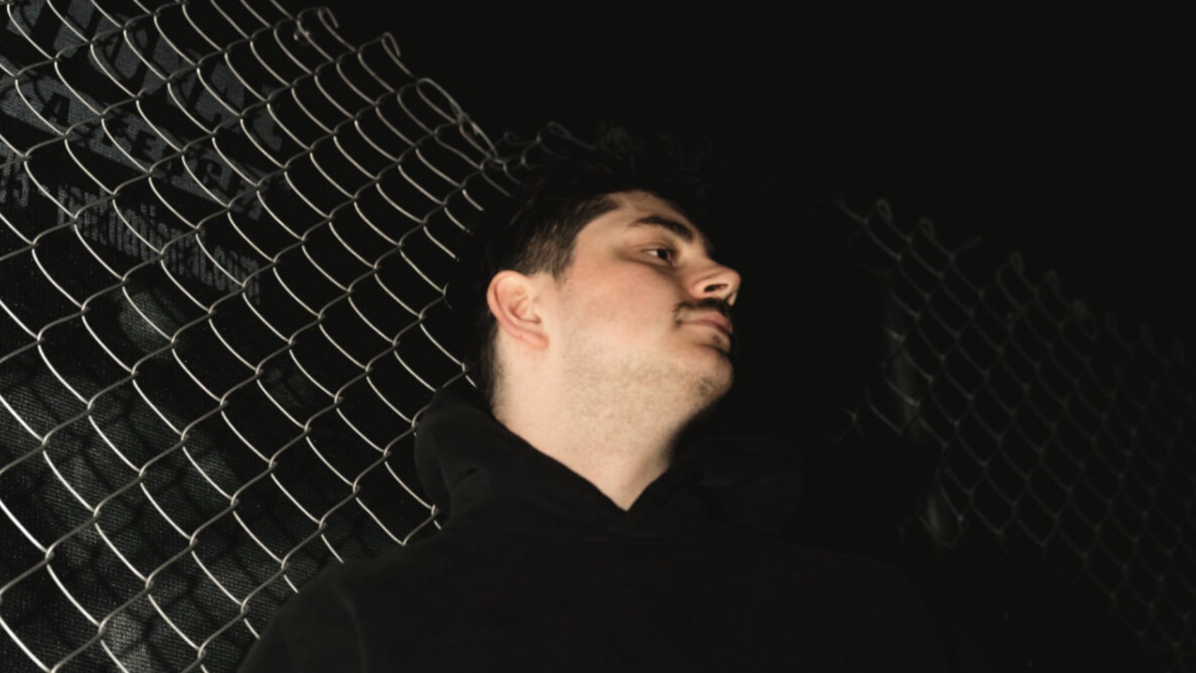 H4RRIS, EDM producer Max Harris, lit against a chain-link fence at night with his head tilted back