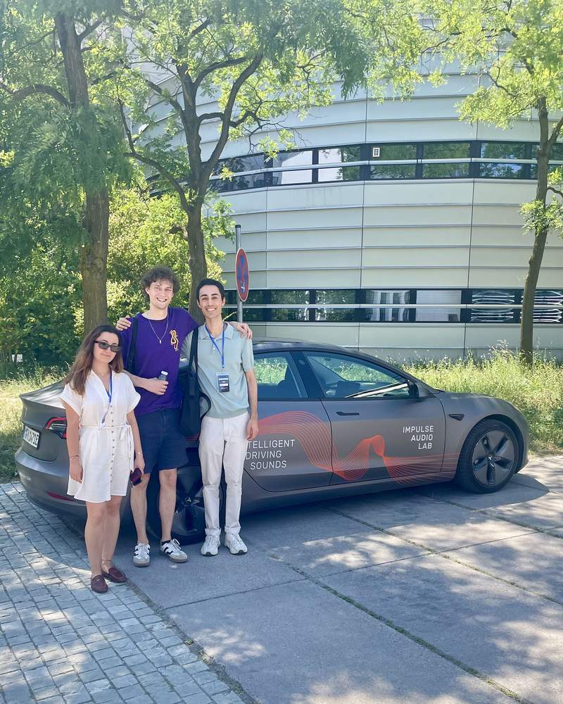 Three Hackatune 2026 participants pose beside Impulse Audio Lab's Intelligent Driving Sounds test car
