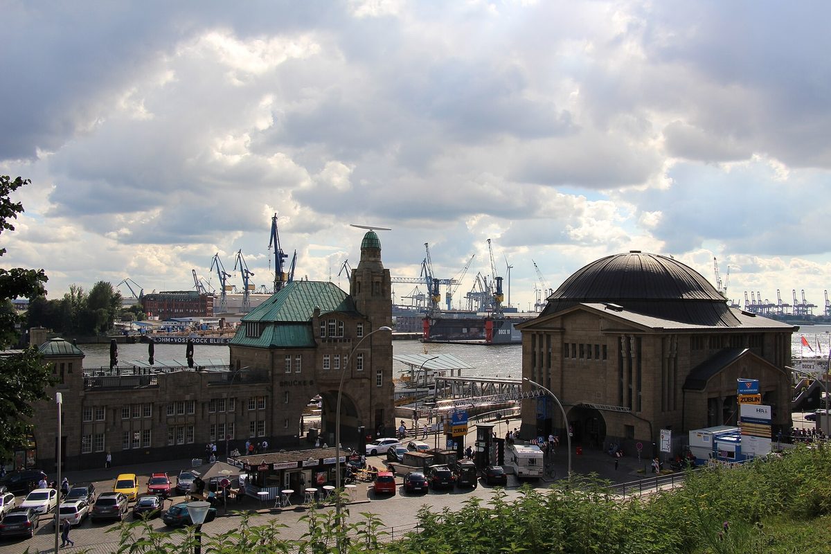 The St Pauli Landungsbrucken pier buildings in Hamburg with the harbour cranes behind them under a cloudy sky.