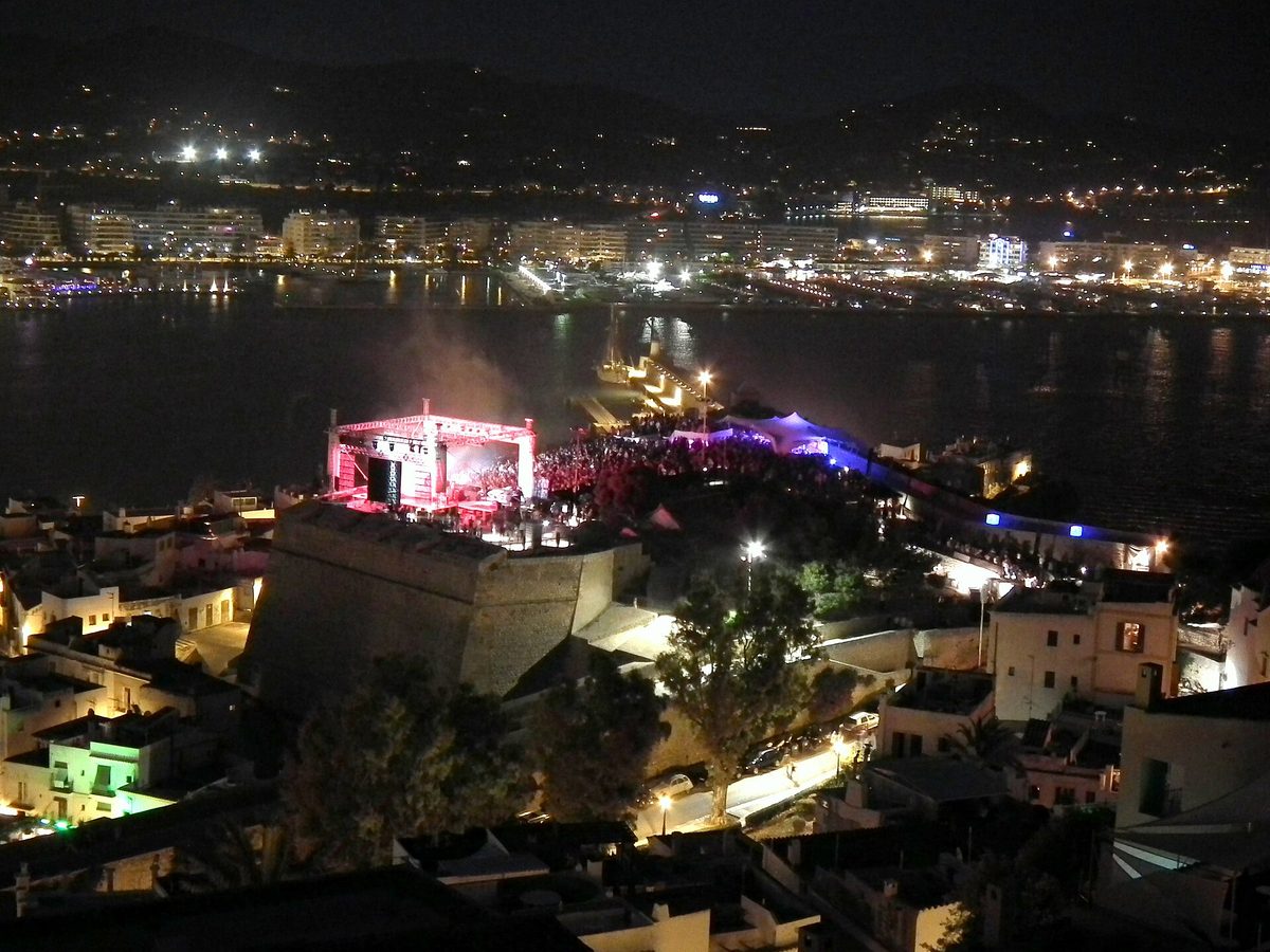 A lit stage and crowd inside the fortified walls of Dalt Vila at night, with the lights of Ibiza town and the bay spreading out behind.