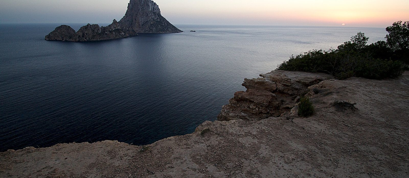 The rock island of Es Vedra rising out of a calm sea off the Ibiza coast at dusk, seen from a scrubby clifftop with the sky fading to pale gold.