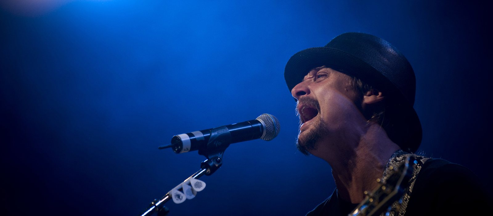 Kid Rock singing into a microphone under blue stage lights in 2008, years before he recorded Halfway 2 Jesus