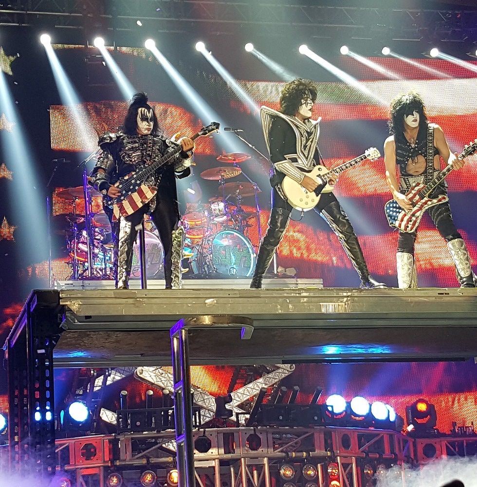 KISS performing live in full makeup on a raised stage in front of an American flag backdrop