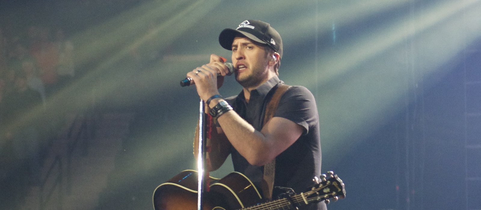 Luke Bryan singing into a microphone on stage with an acoustic guitar during a 2014 concert