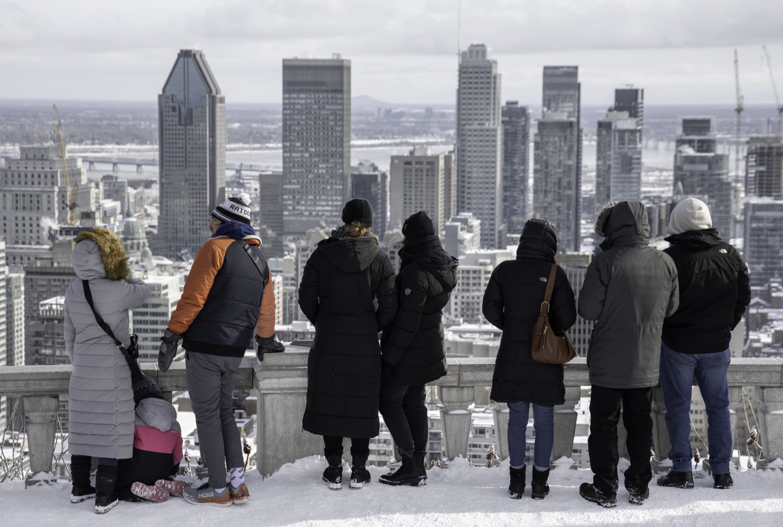 Visitors in winter coats at the Kondiaronk Belvedere on Mount Royal looking out over the snow-covered downtown Montreal skyline.