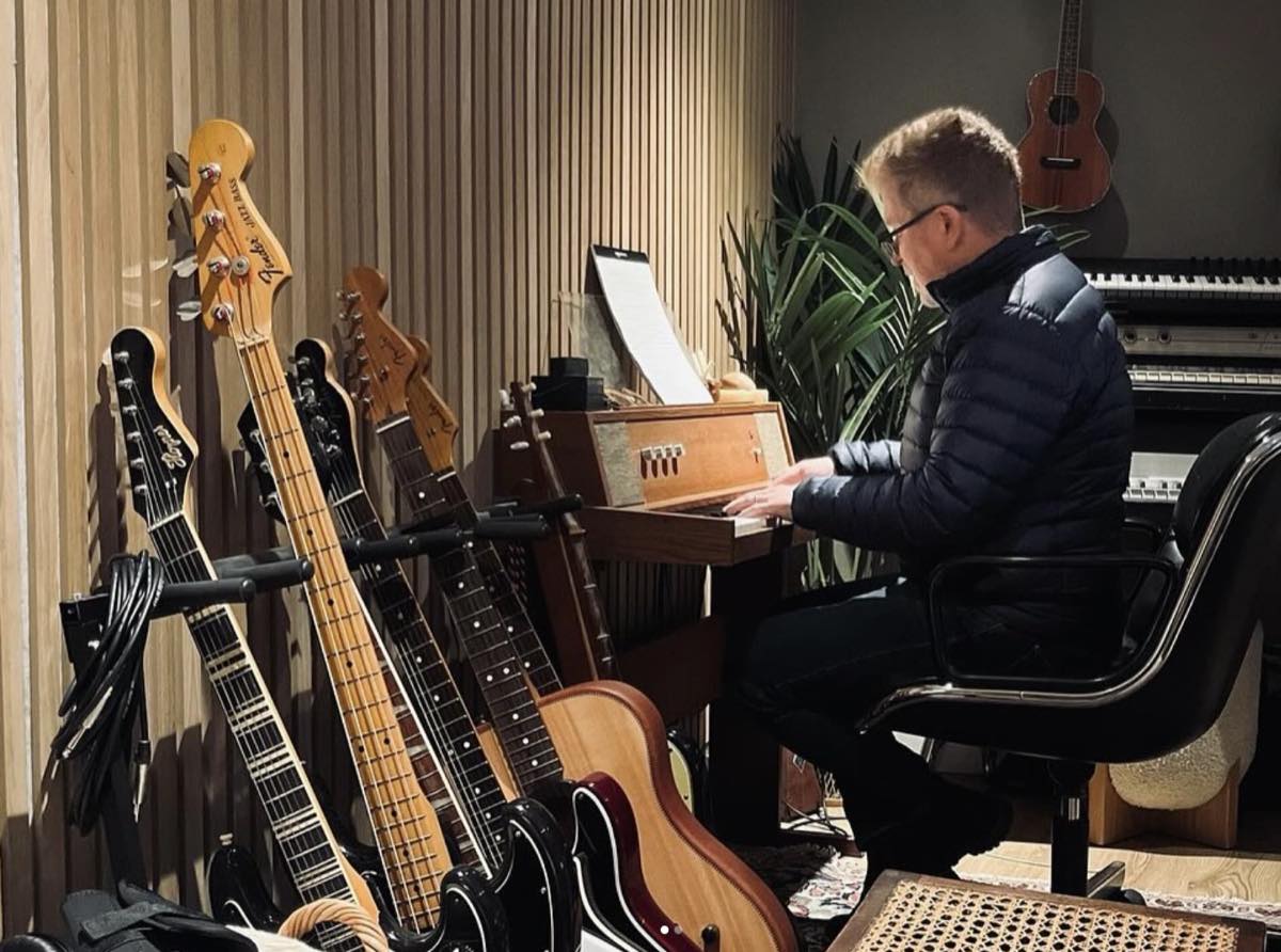 Producer Matt Rollings plays a harmonium in the studio beside a rack of electric guitars and a piano
