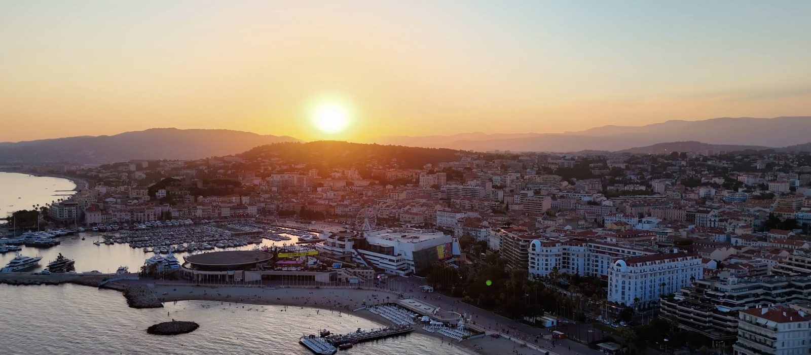 Cannes seen from above at sunset, the bay and marina in the foreground and the town spreading back towards hills under an orange sky.