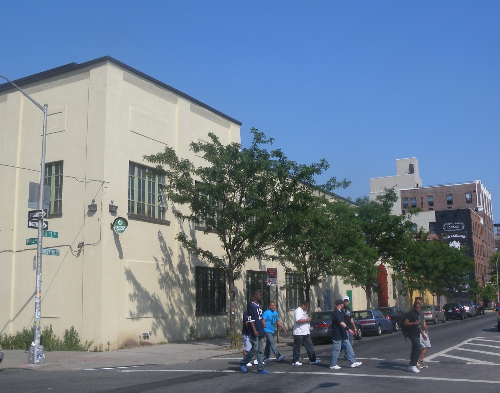 The corner of Wythe Avenue and North 11th Street in Williamsburg, Brooklyn, with the low brick Brewers Row buildings along the block and people crossing the street in summer.