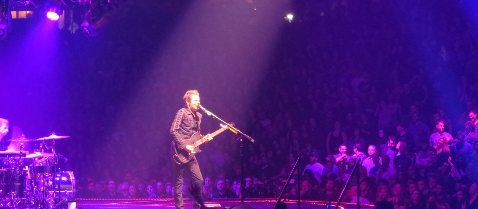Muse frontman Matt Bellamy singing and playing guitar on a lit arena stage in front of a packed crowd