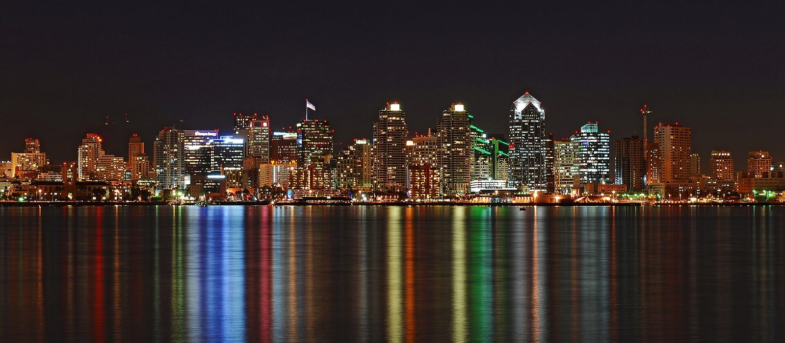 The downtown San Diego skyline lit at night, its towers reflected in long coloured streaks across the calm water of the bay.