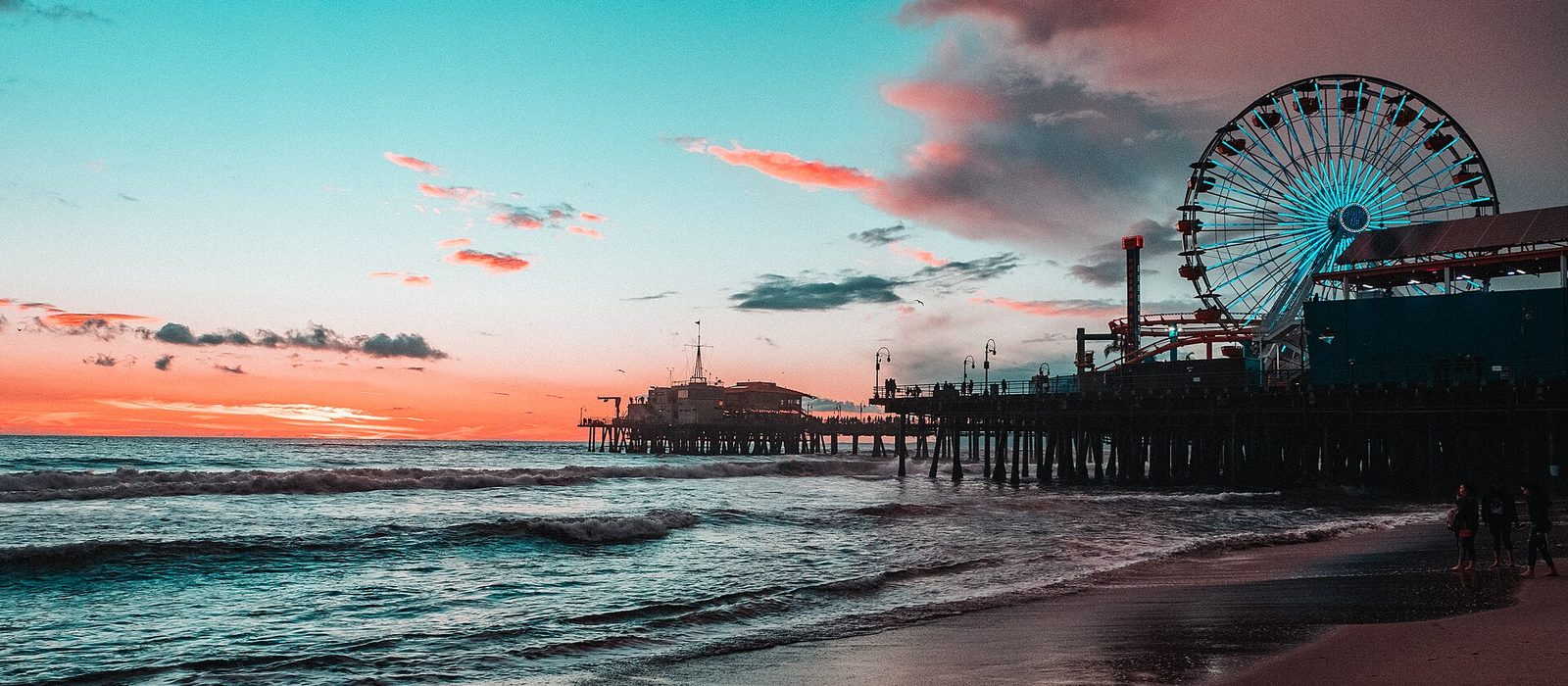 Santa Monica Pier at sunset seen from the beach, its Ferris wheel and rollercoaster lit against a pink and turquoise sky with waves in the foreground.