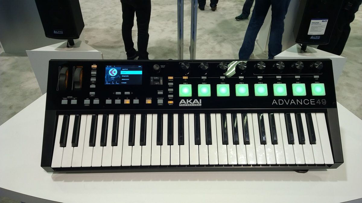 An Akai Professional Advance 49 keyboard controller on a white plinth at the NAMM Show, its screen showing Logic Pro X, Bitwig Studio and Ableton Live in a browser list, with Alto speakers either side.