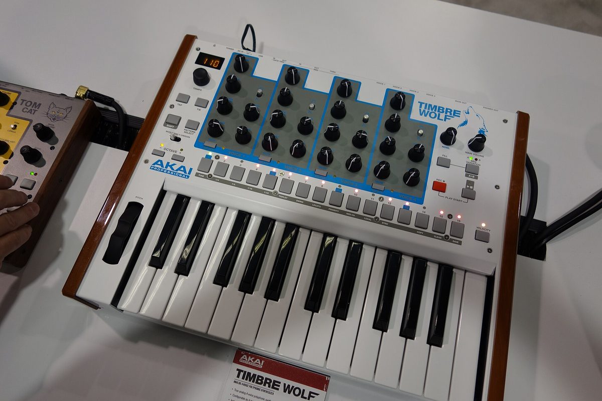 An Akai Professional Timbre Wolf analogue polyphonic synthesizer on a white display table at the NAMM Show, with a hand reaching for an Akai Tom Cat drum machine beside it.