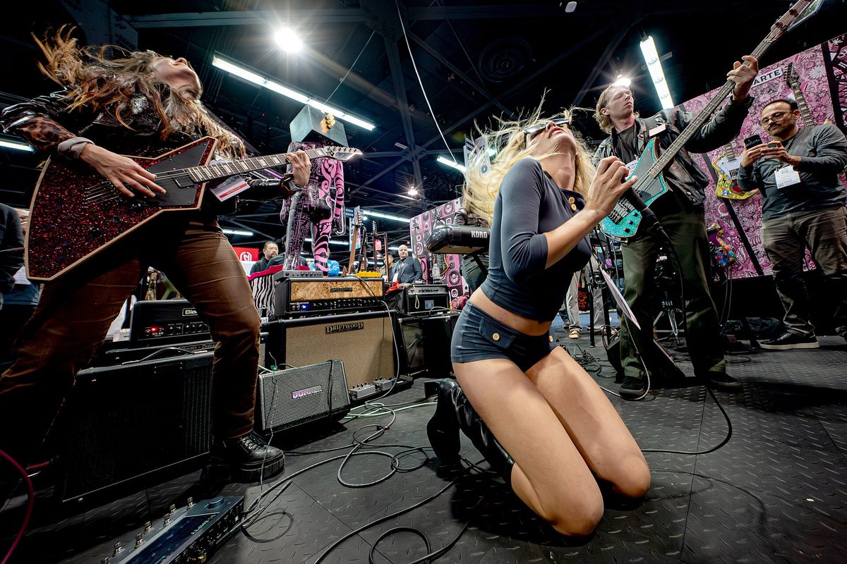 Three musicians playing electric guitars and bass at full tilt on a guitar booth at the NAMM Show, surrounded by amplifiers and attendees filming on phones.