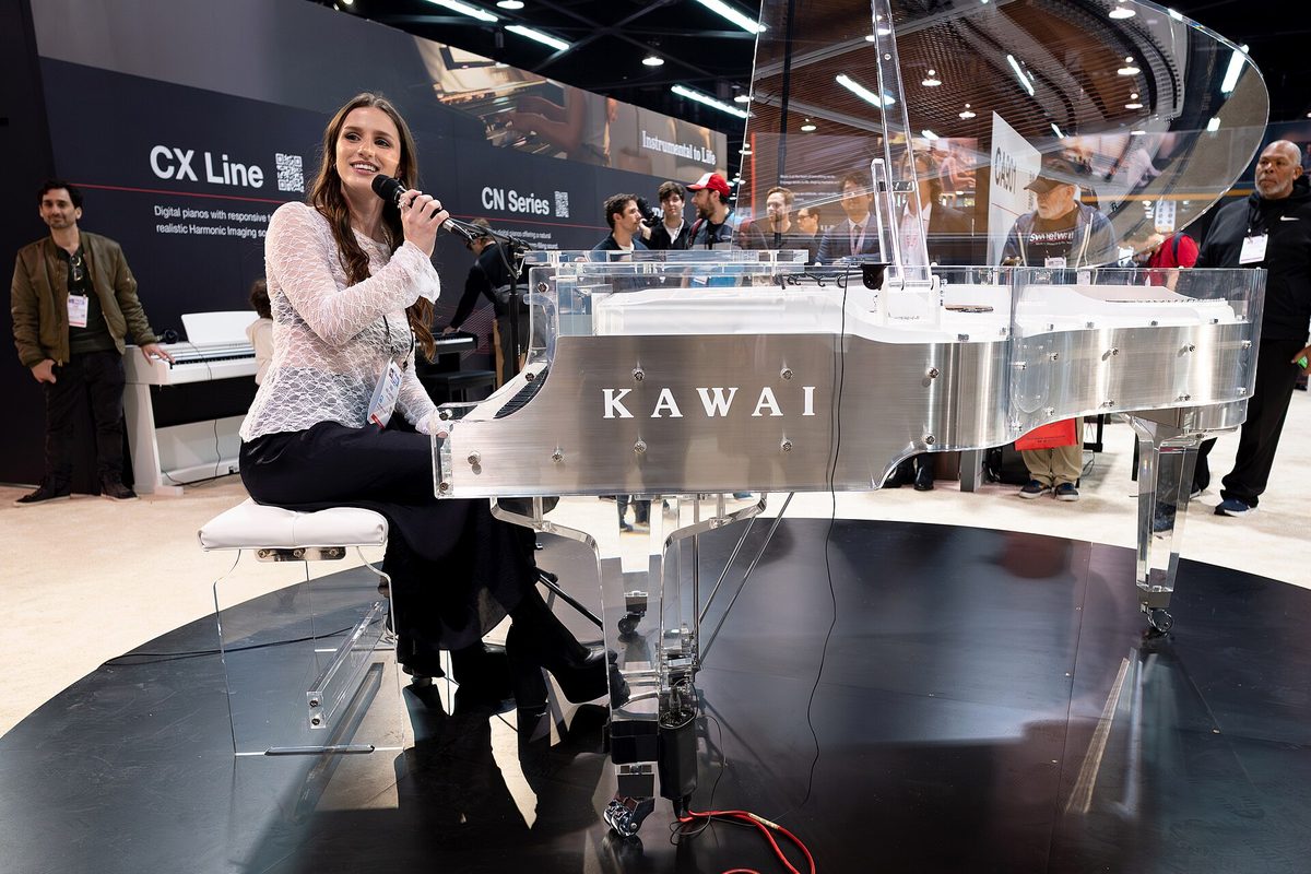 A singer at a transparent Kawai grand piano on the Kawai booth at the NAMM Show, with CX Line and CN Series signage behind and badged attendees watching.
