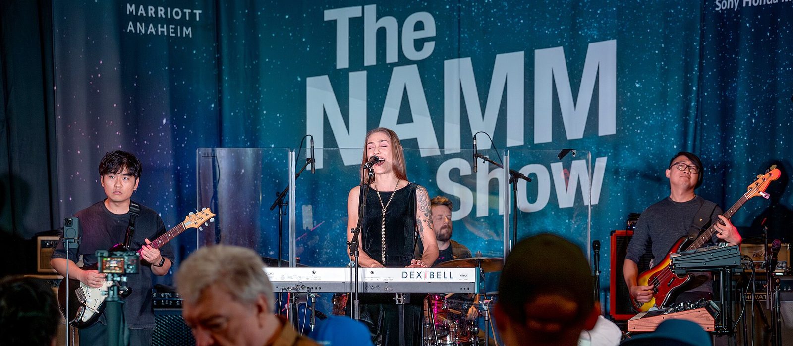 A band performing on a stage in front of a large starfield backdrop reading The NAMM Show, with a Dexibell keyboard in front and a crowd of badged attendees watching.