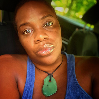 Olivia B. Moore — selfie in a car, wearing a blue tank top and a polished green stone pendant necklace, with glossy lipstick and natural light through the rear window.
