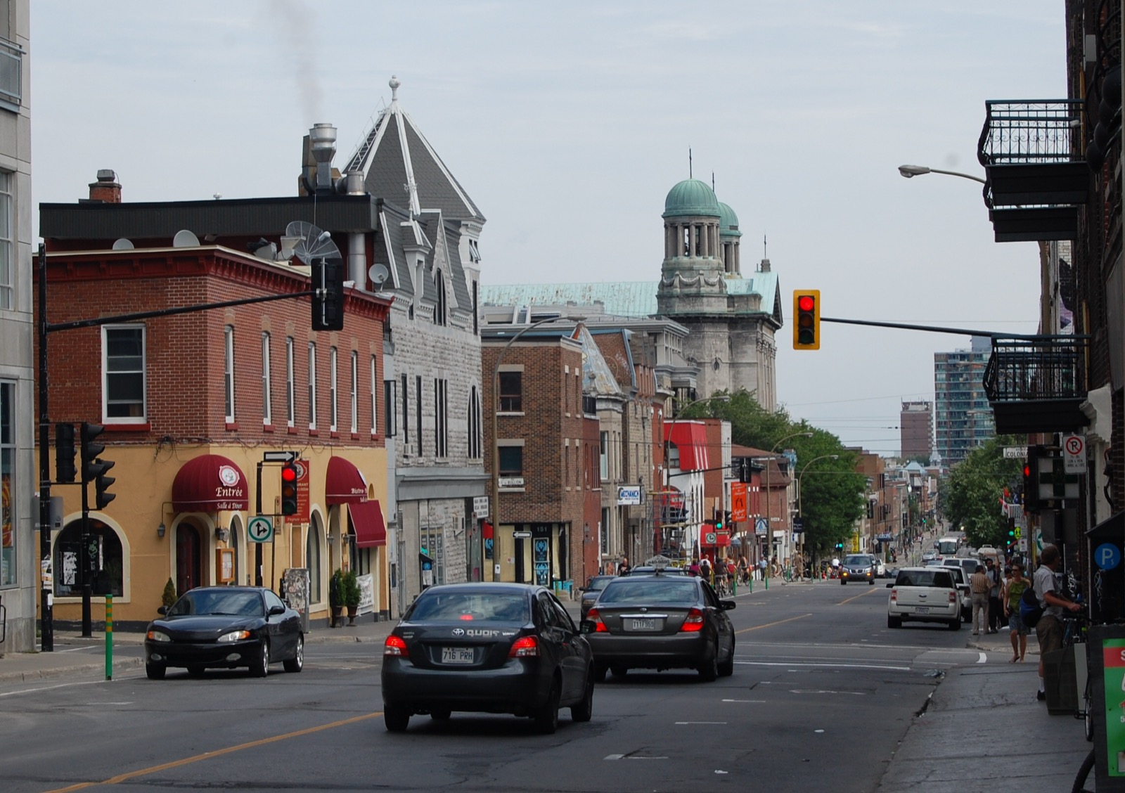 A busy Montreal commercial street in the Plateau with bistros, red awnings and a green copper dome rising behind the rooftops.