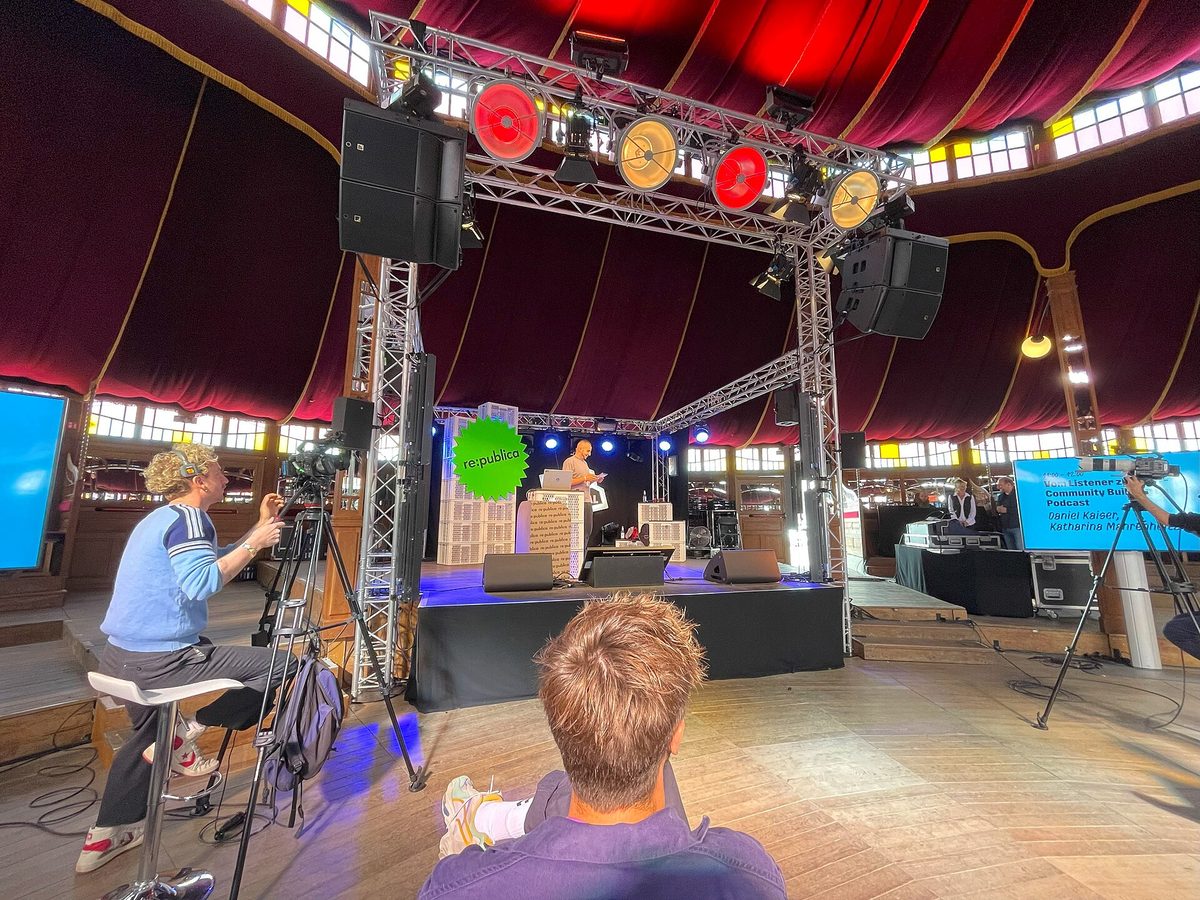 A conference stage under a red circus-tent ceiling with a truss lighting rig, screens and camera operators filming a session in progress.
