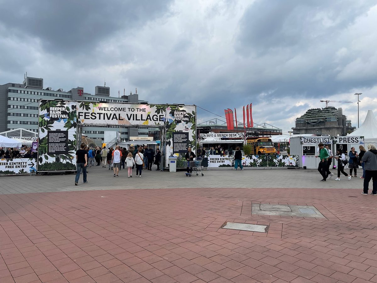 The entrance to the Reeperbahn Festival Village on Heiligengeistfeld in Hamburg, with a Welcome to the Festival Village banner, stalls and a crowd on the paved square.