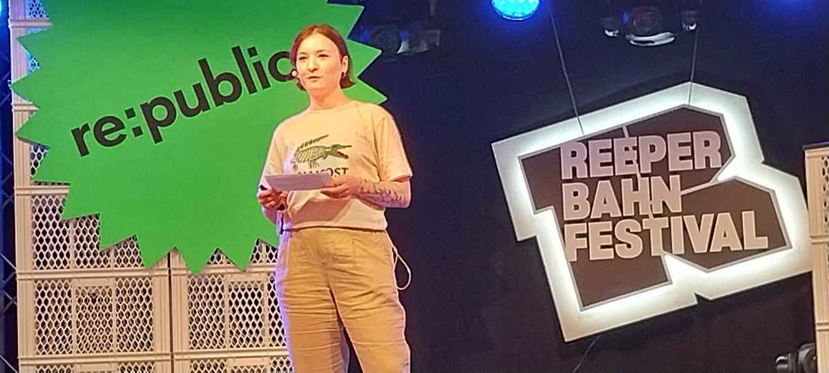 A speaker standing at a lectern on a conference stage between a large green re:publica logo and an illuminated Reeperbahn Festival sign.