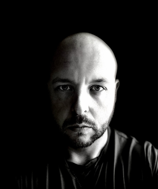 Black-and-white portrait of a bald, bearded man against a black background, used as the Rebel Redeemed 316 artist image.