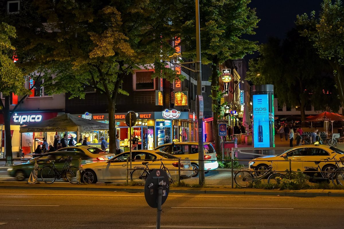 The Reeperbahn at dusk with lit shopfronts and betting-shop signage, cars and taxis queueing along the wide boulevard under plane trees.