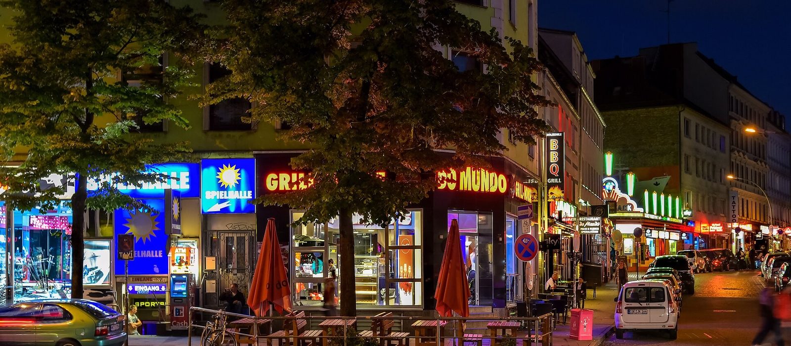 The Reeperbahn in Hamburg at night, lined with neon signs for amusement arcades, bars and snack stands, with pavement tables under trees and traffic passing.