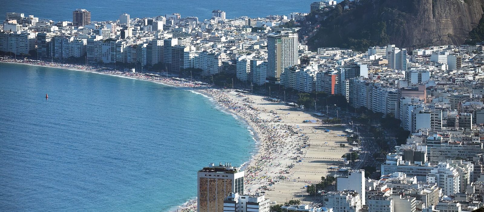 The curve of Copacabana beach in Rio de Janeiro seen from above, pale sand meeting turquoise water with the dense white apartment blocks of the neighbourhood behind it.